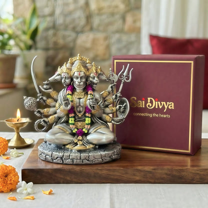 A silver and gold-toned statue of the Hindu goddess Sri Divya seated on a stone base, displayed next to a dark red gift box with gold trim and the words 'Sri Divya connecting the hearts.' The scene is styled on a wooden surface with a lit brass candle, orange marigold flowers, and a softly blurred indoor background.